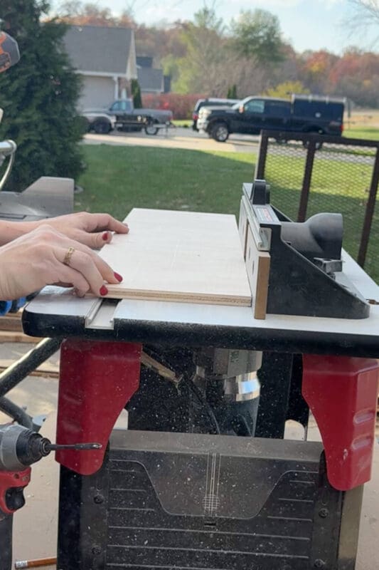 Close-up of plywood sliding across a router table to cut grooves for a DIY kitchen drawer organizer.