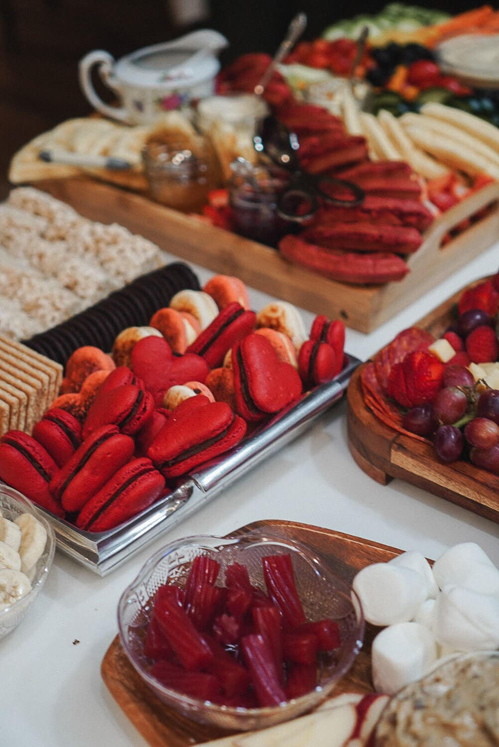 Dessert tray at a Galentine's Gathering with bright red heart macarons, Oreos, graham crackers, red licorice bites, and marshmallows, with fruit and waffles in the background.