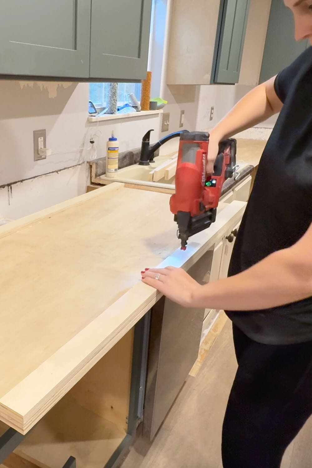 Using a red brad nailer to secure the front trim piece onto plywood DIY countertops, creating a thicker countertop edge before adding faux marble and epoxy.