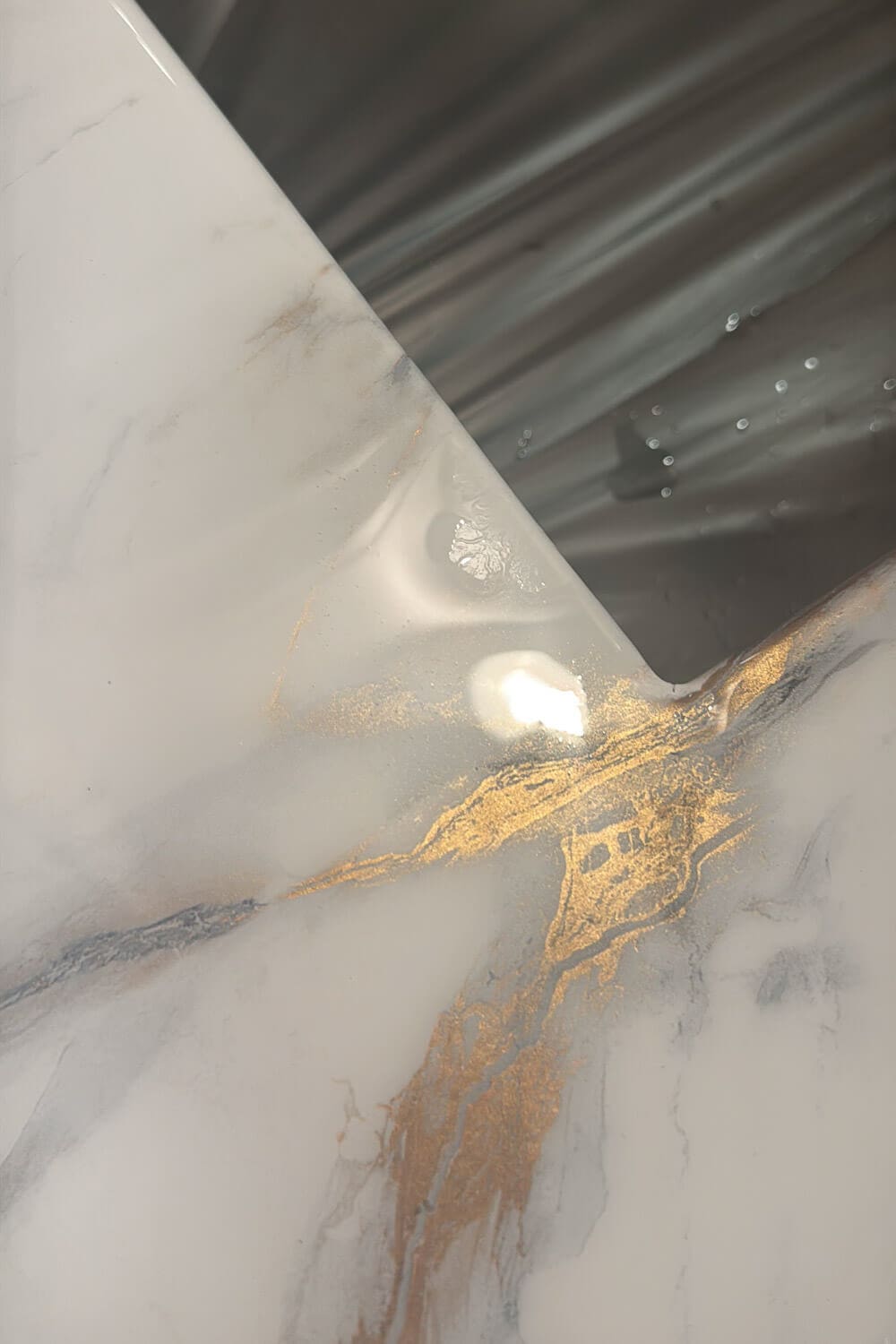 Close up of DIY countertops that look like marble, showing glossy epoxy over marble contact paper with metallic gold veining at the corner of a stainless steel sink.