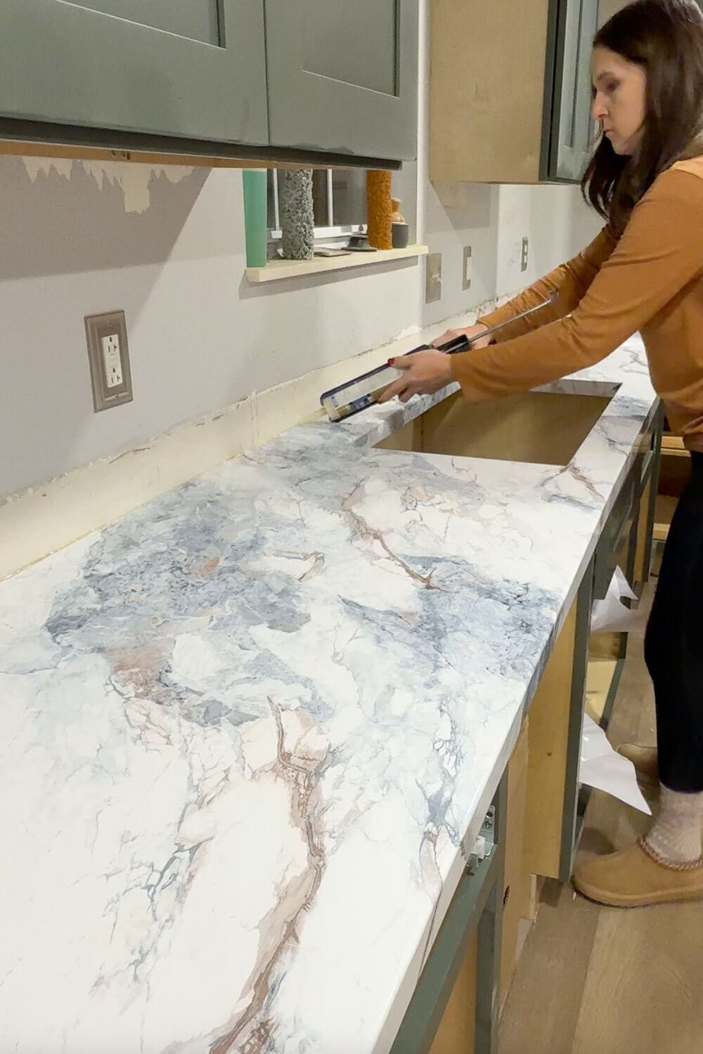 Woman installing marble contact paper on new DIY countertops, smoothing the faux marble finish across a long plywood kitchen counter.