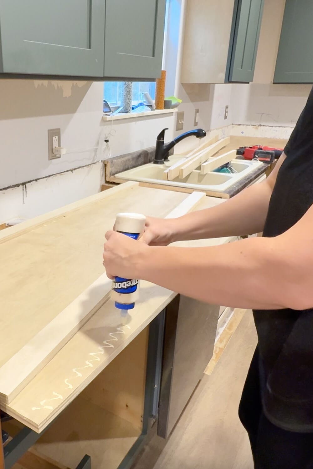 Applying wood glue to the front edge of plywood while building DIY countertops, prepping the raw kitchen cabinets for new plywood countertop installation.