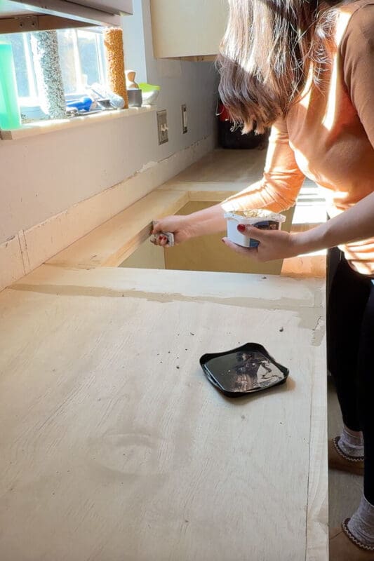 DIYer applying wood filler to the front edge of new plywood kitchen countertops as a first step before installing faux marble epoxy DIY countertops.