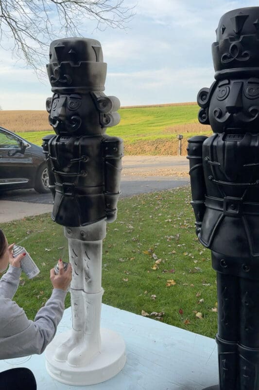 Woman spray painting the legs of a giant nutcracker while another black giant nutcracker stands finished beside it in the yard.