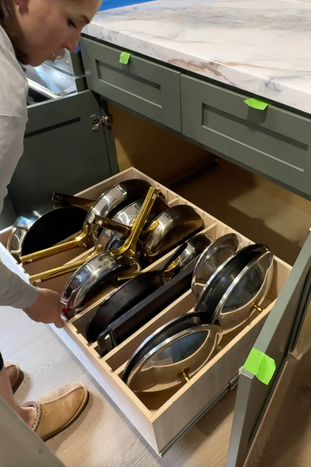 Woman pulling out a custom slide-out drawer filled with neatly organized pots, pans, and lids in vertical slots, showing the finished DIY pots and pans storage organizer inside a lower kitchen cabinet.