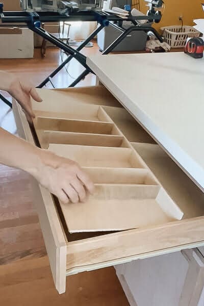 Hands sliding a custom plywood utensil drawer organizer with divided compartments into a shallow kitchen drawer for neatly sorted cooking tools.