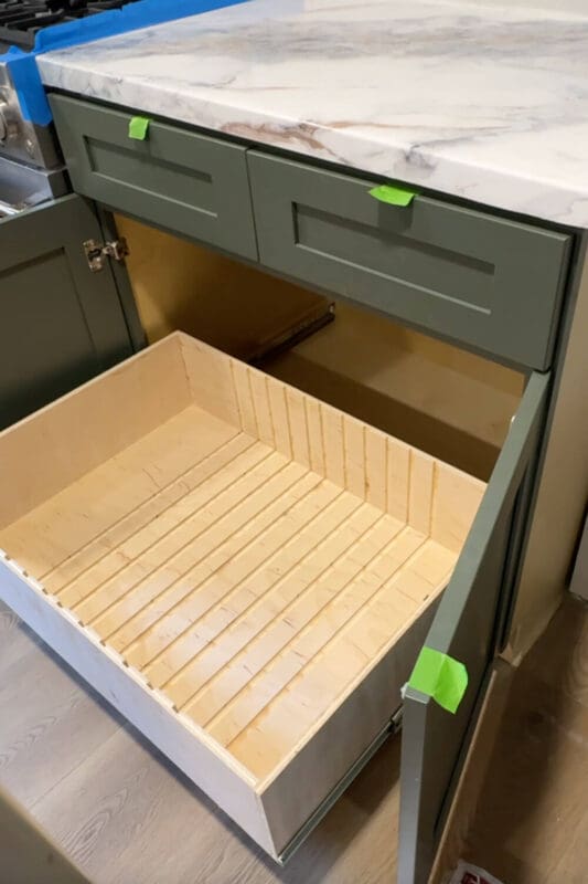 Empty slide-out plywood drawer installed in a green lower cabinet, showing evenly spaced grooves ready for adjustable dividers and custom pots and pans storage.