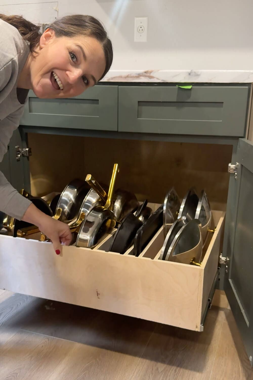 Smiling woman kneeling beside an open lower cabinet, proudly showing a fully loaded slide-out drawer with organized pots, pans, and lids in custom dividers for easy pots and pans storage.