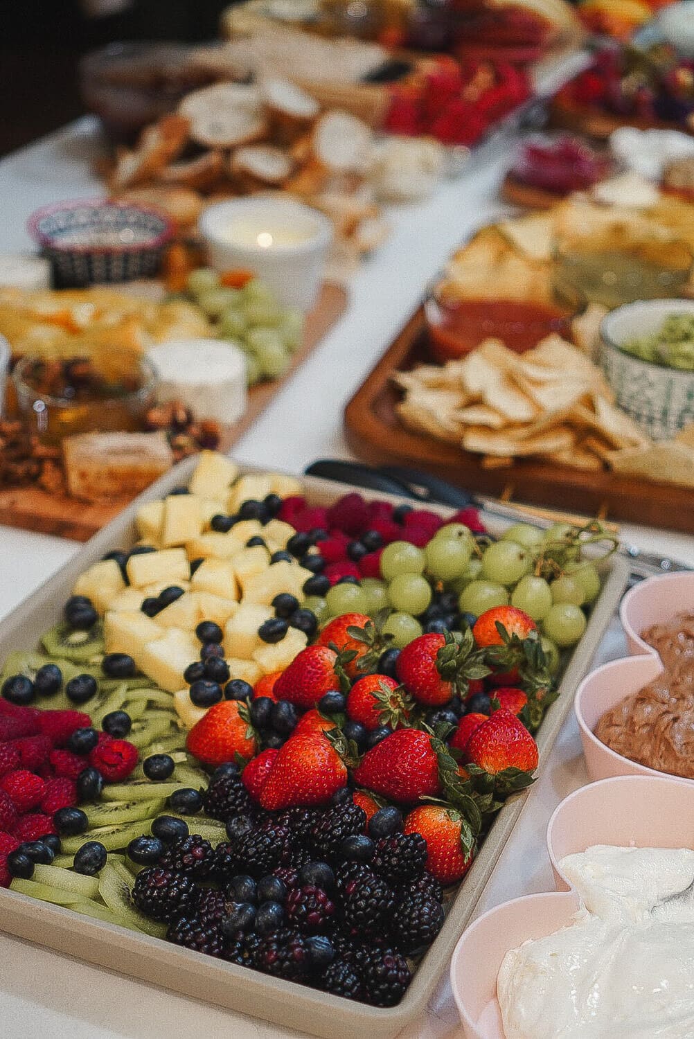 Large fruit tray with strawberries, blackberries, blueberries, kiwi, raspberries, pineapple, and grapes beside heart-shaped bowls of marshmallow fluff and chocolate fruit dip at a charcuterie party.