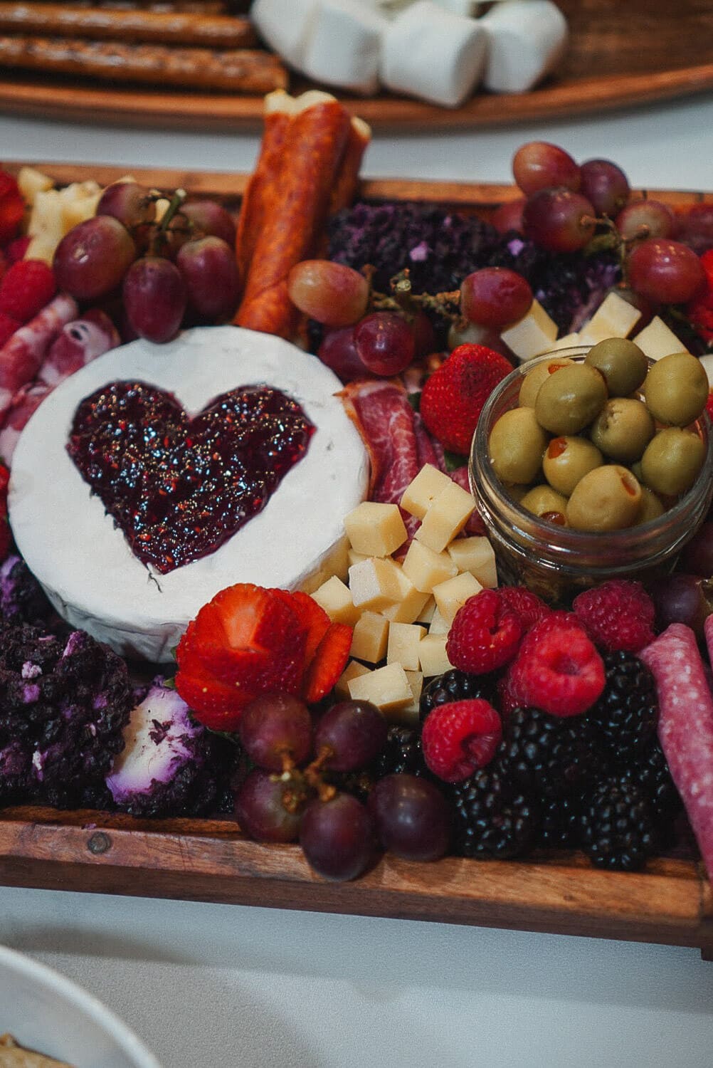 Galentine’s charcuterie party board with brie topped with a jam heart, grapes, olives, cheese cubes, raspberries, blackberries, and salami—cute, shareable appetizer spread.