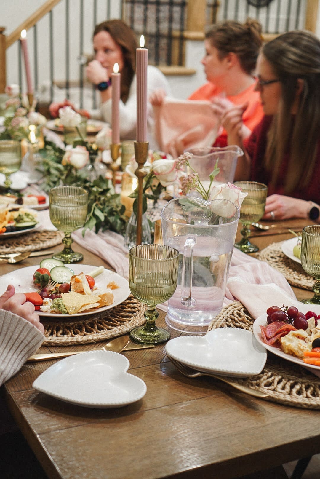 Galentine’s Day tablescape with pink taper candles, green glass goblets, woven chargers, and heart-shaped plates as friends enjoy food and desserts.