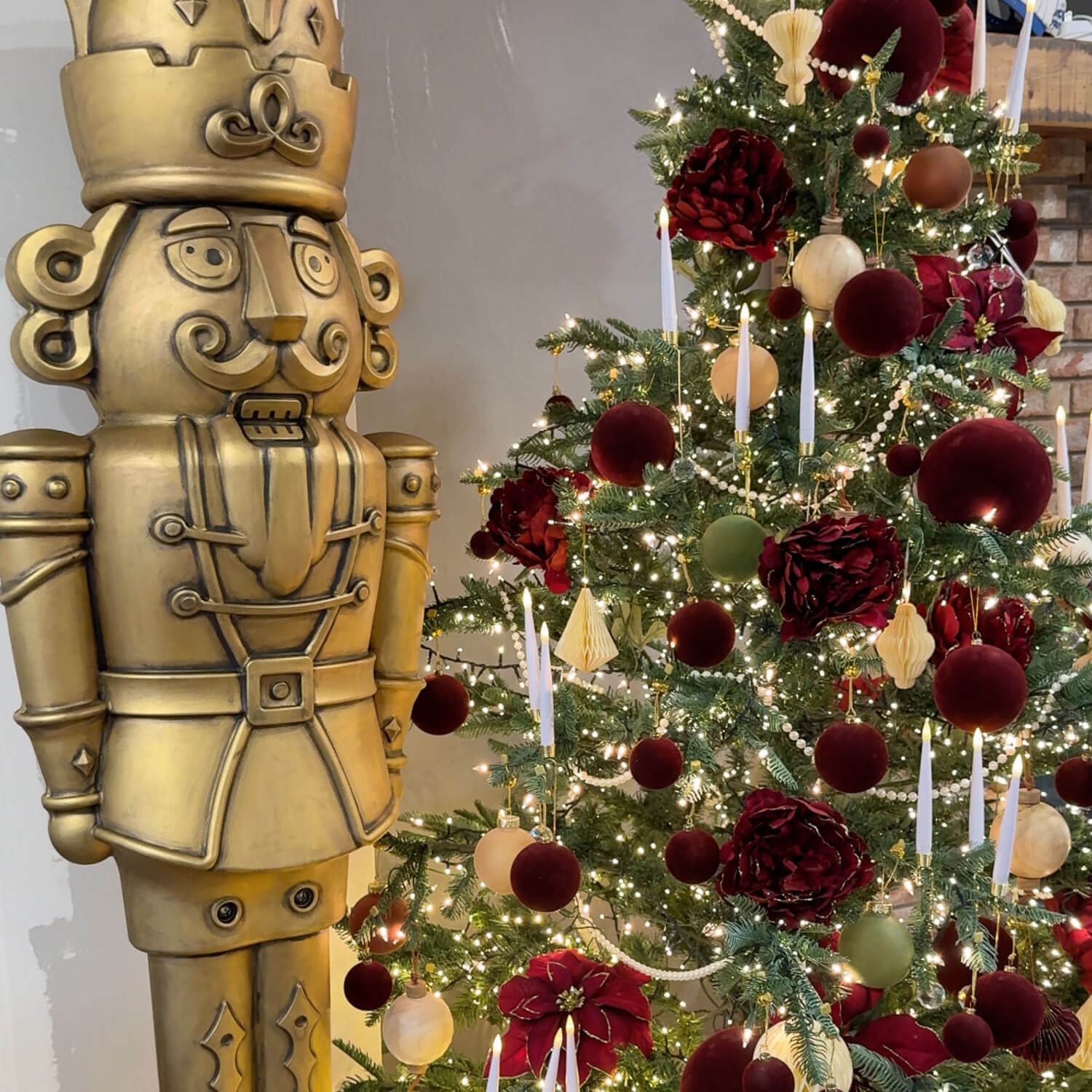 Antique bronze giant nutcracker standing beside a Christmas tree decorated with burgundy, green, and neutral ornaments and glowing candles.