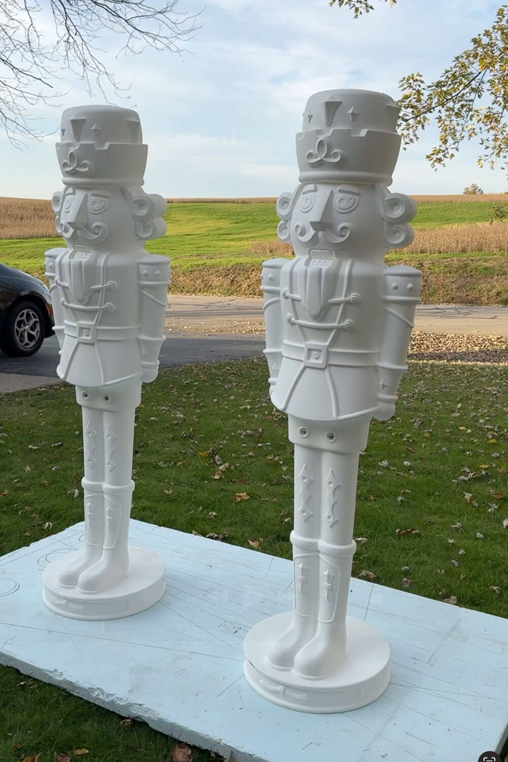Two tall white giant nutcrackers standing outside on a sheet of foam board before being painted for a DIY Christmas decor project.