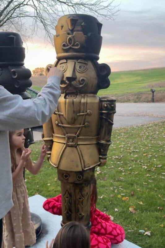 Mom and kids adding final Rub n Buff details to a giant bronze nutcracker during a family DIY Christmas project in the yard.