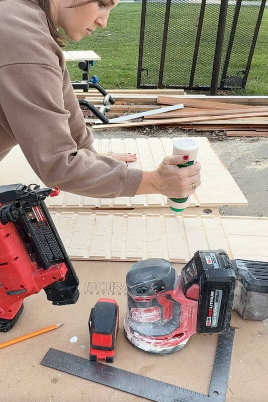 Woman applying wood glue to routed grooves on a plywood panel while building a DIY slide-out drawer for custom pots and pans storage, with nail gun and sander on the workbench.