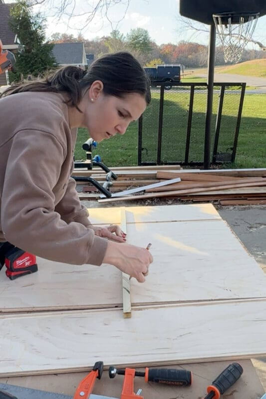Woman using a wood spacer to mark evenly spaced lines on plywood, laying out the vertical dividers for a custom pots and pans storage organizer.