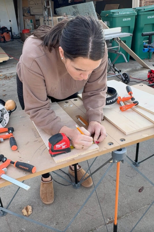 Woman marking measurements on a plywood board at an outdoor workbench, laying out where the grooves will be cut so the interlocking drawer pieces for the organizer slide together.