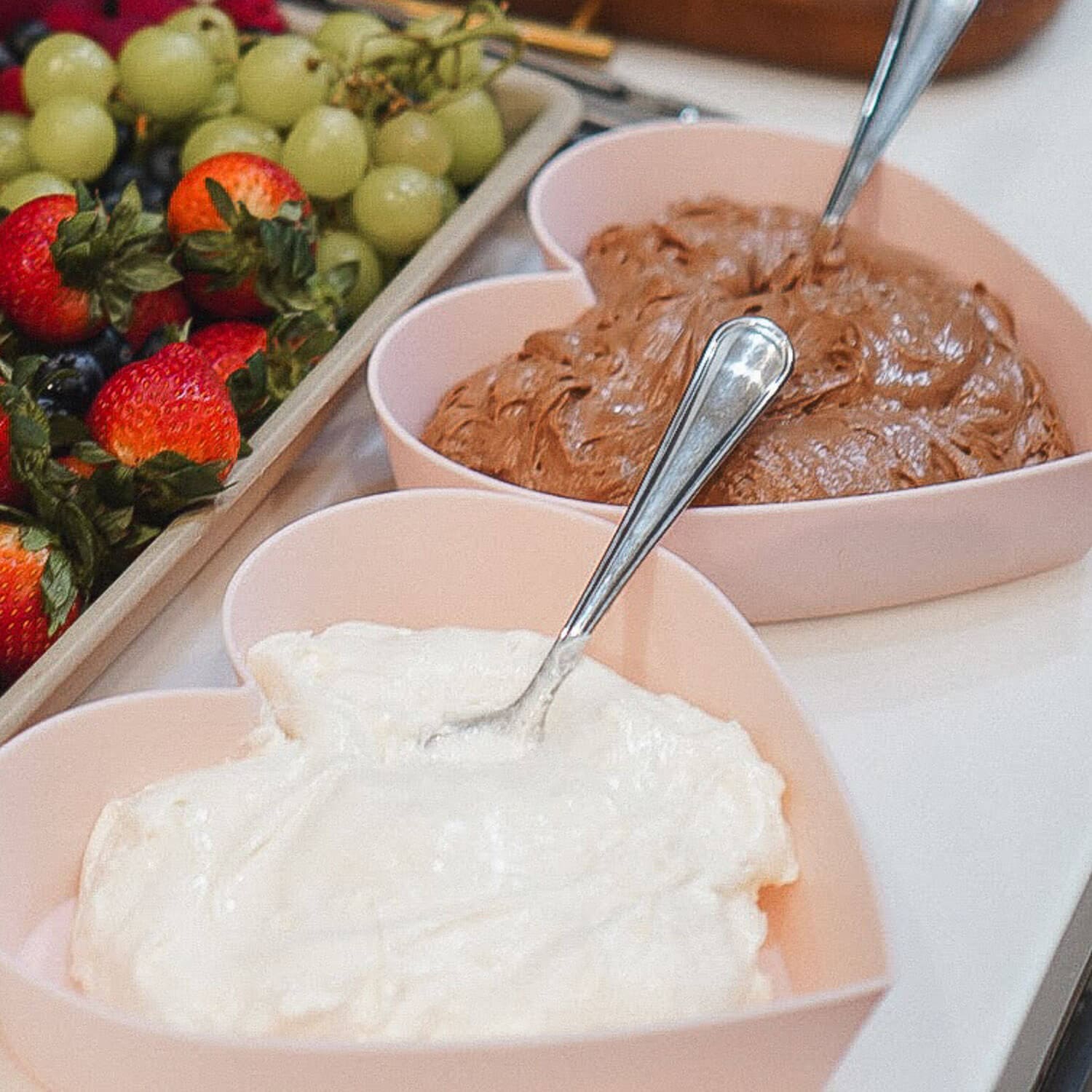 Heart-shaped bowls of marshmallow fluff fruit dip and chocolate fruit dip with spoons beside a fruit tray of strawberries, grapes, and blueberries.
