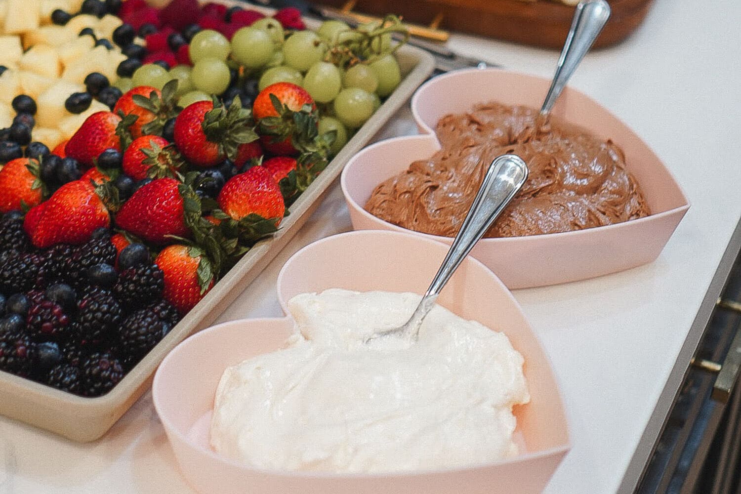Heart-shaped bowls of marshmallow fluff fruit dip and chocolate fruit dip with spoons next to a colorful fruit tray of strawberries, grapes, blueberries, and blackberries.