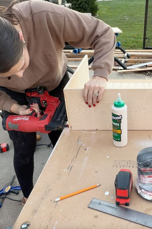 Woman using a cordless brad nailer to attach the side of a plywood drawer box, assembling a sturdy slide-out cabinet for organized pots and pans storage.