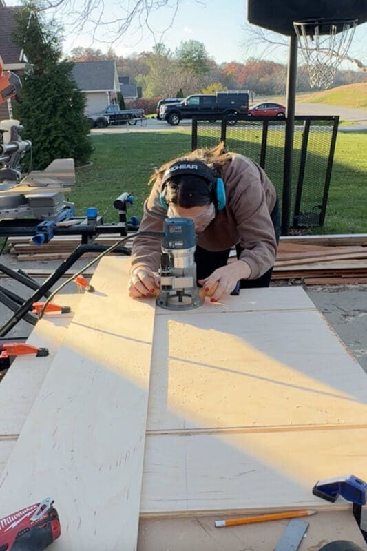 Woman wearing ear protection and safety glasses using a handheld router along a straightedge to cut long grooves in plywood panels for a DIY slide-out pots and pans storage drawer.