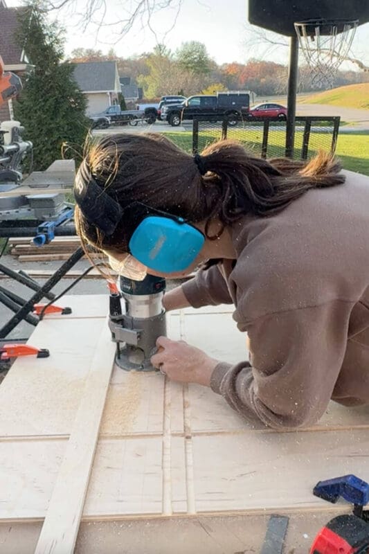 Side view of woman routing multiple parallel grooves in plywood with a guide board clamped in place, creating slots for the vertical dividers in a custom pots and pans storage organizer.