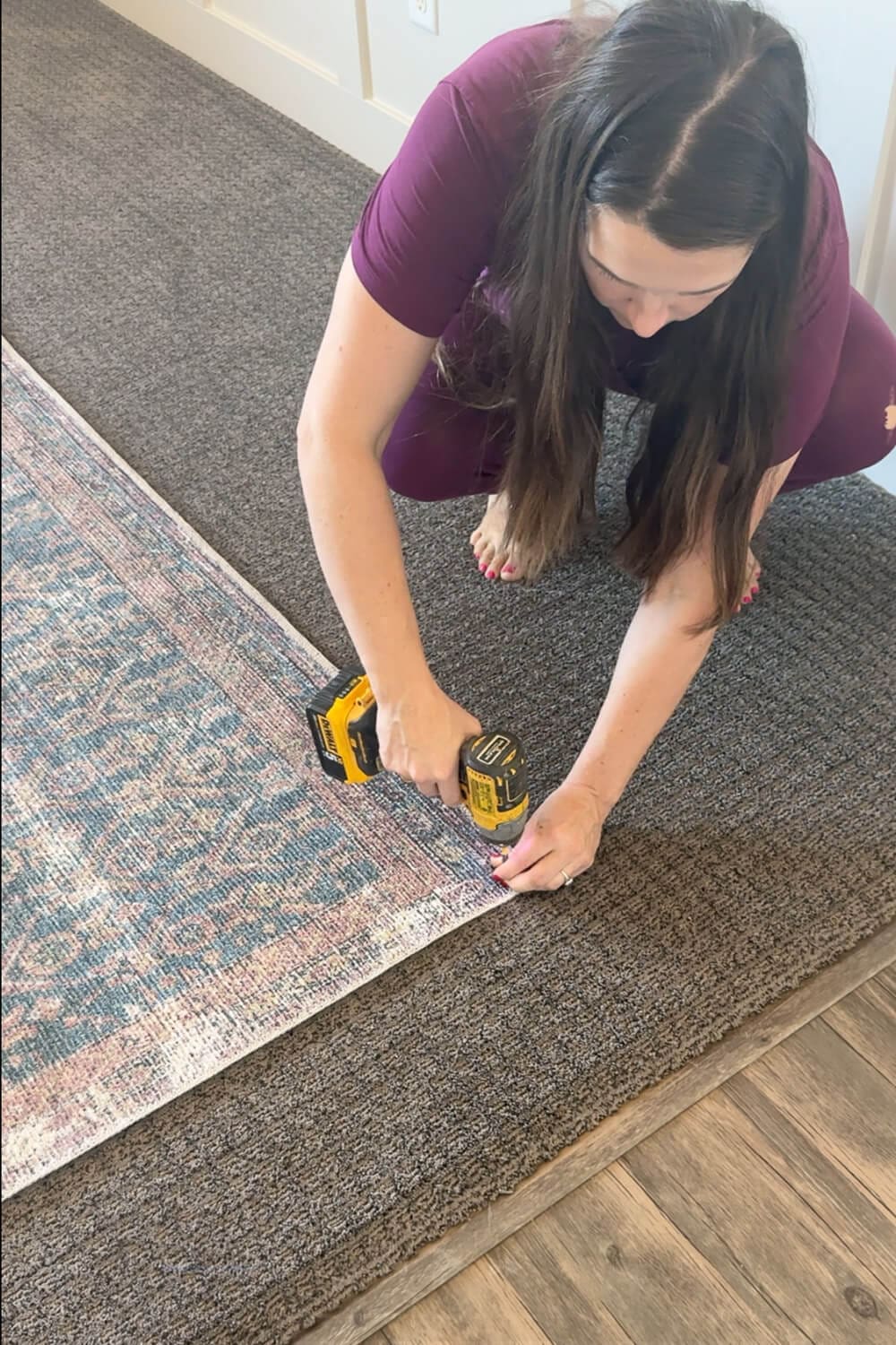 A woman using a drill to screw the corner of an area rug into the carpeted floor to stop sliding during a living room makeover. Works way better than rug tape.