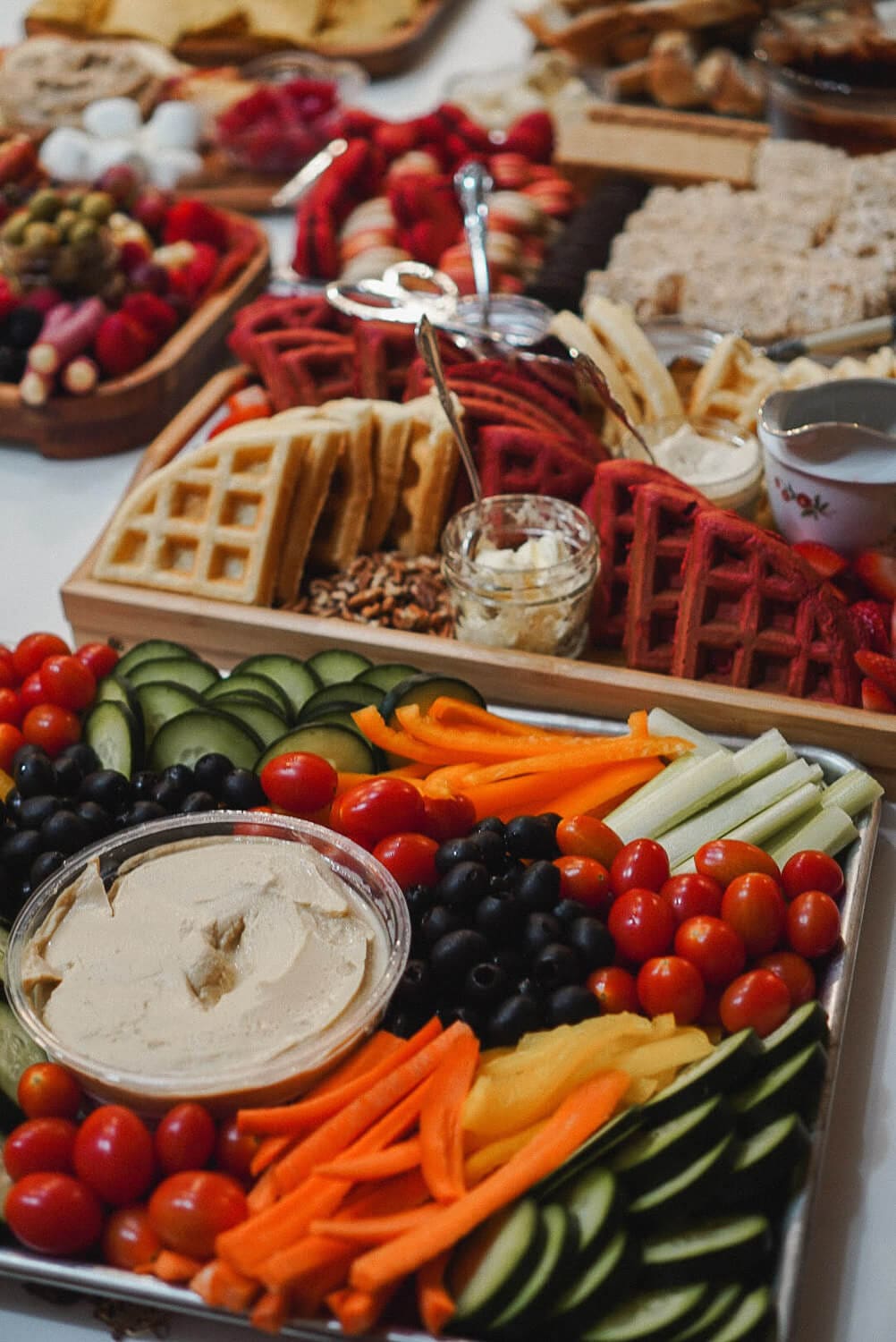 Veggie charcuterie board for a party with hummus, cucumbers, cherry tomatoes, black olives, carrots, celery, and bell peppers; waffle brunch board with toppings in the background.