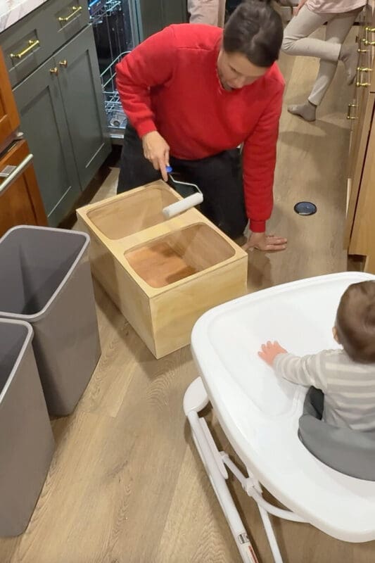 Applying a clear polycrylic topcoat with a foam roller onto a plywood double trash can cabinet insert, sealing the wood for a durable DIY pull out trash can cabinet.