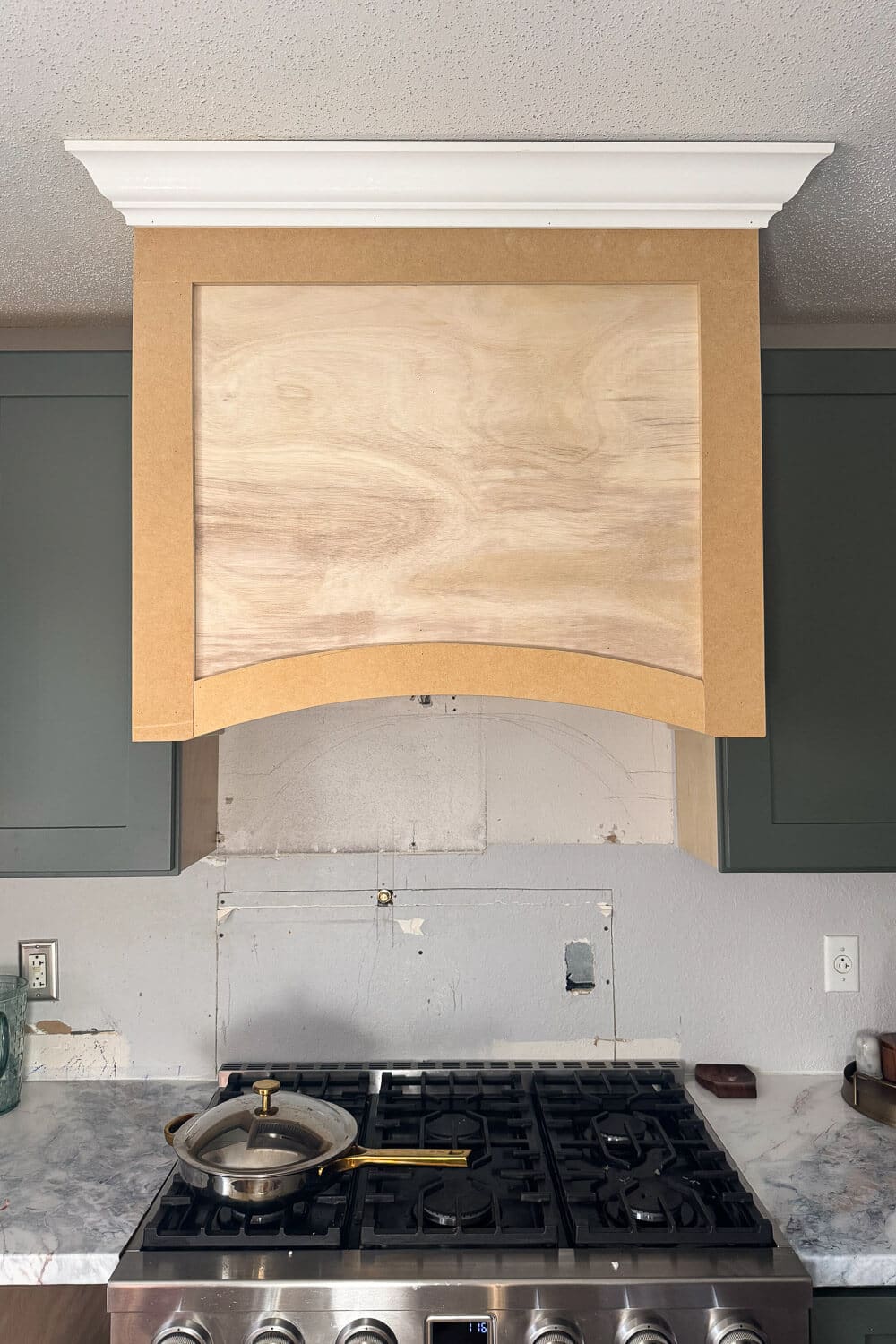 Wide view of an in-progress DIY wood range hood cover framed above a gas stove, featuring an arched bottom trim, plywood center panel, MDF side panels, and white crown molding between dark green kitchen cabinets.
