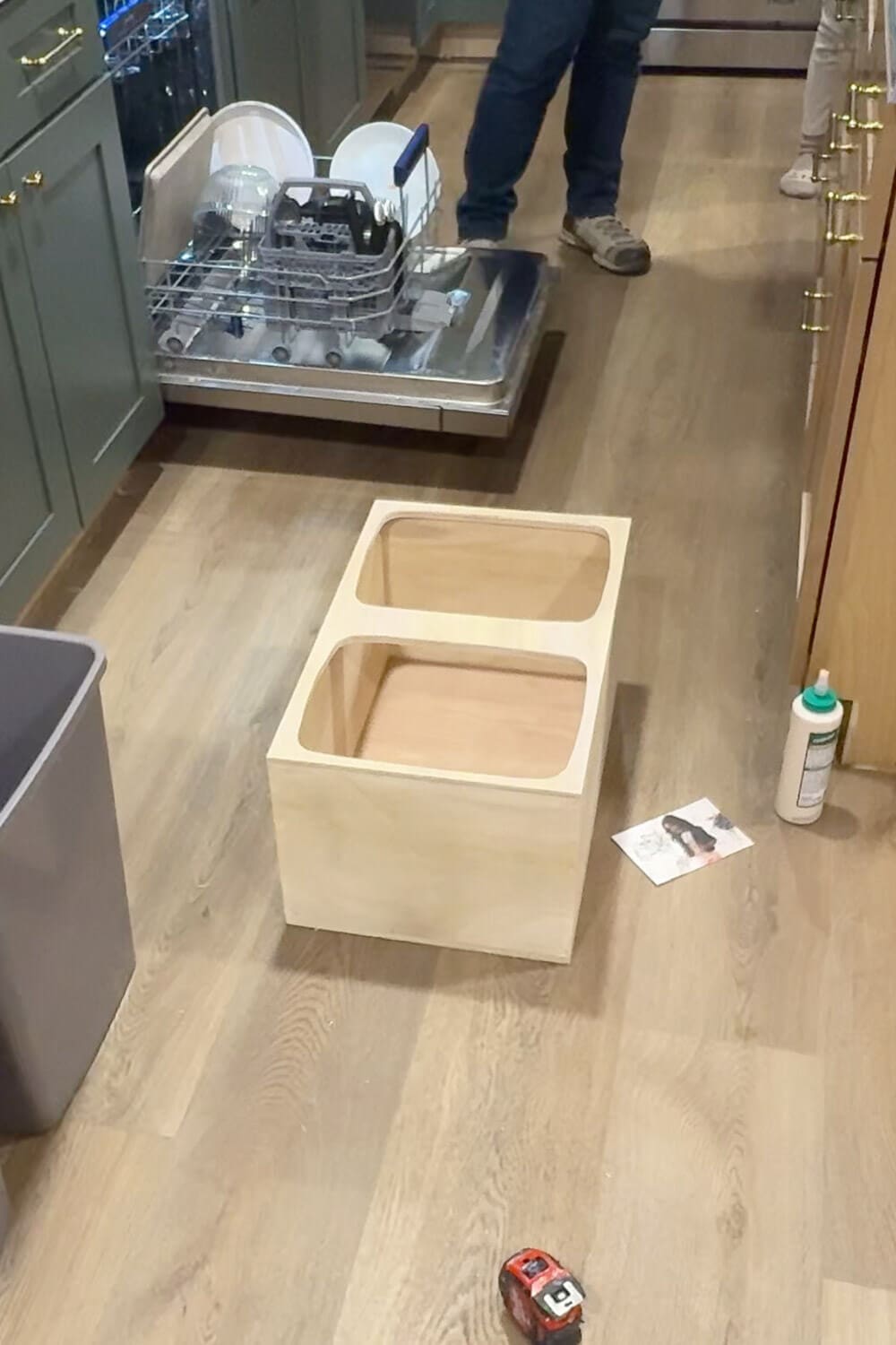 Unfinished plywood box with two rounded trash can openings sitting on the kitchen floor, showing the assembled insert for a DIY pull out double trash can cabinet.
