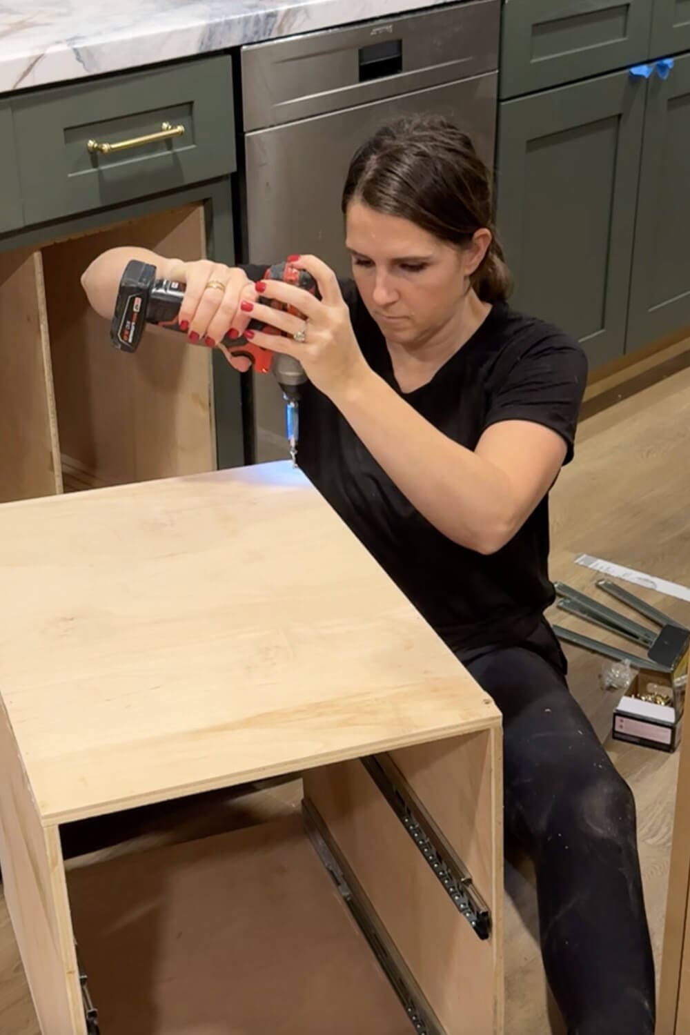 Woman using a drill to screw together a plywood drawer box with side-mounted slides for a DIY pull-out organizer for food storage containers.
