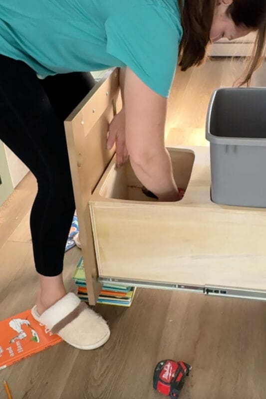 Attaching the cabinet door to the front of the pull out double trash can box while it’s propped up on a stack of kids’ books, aligning the DIY trash can cabinet drawer with the rest of the kitchen cabinets.