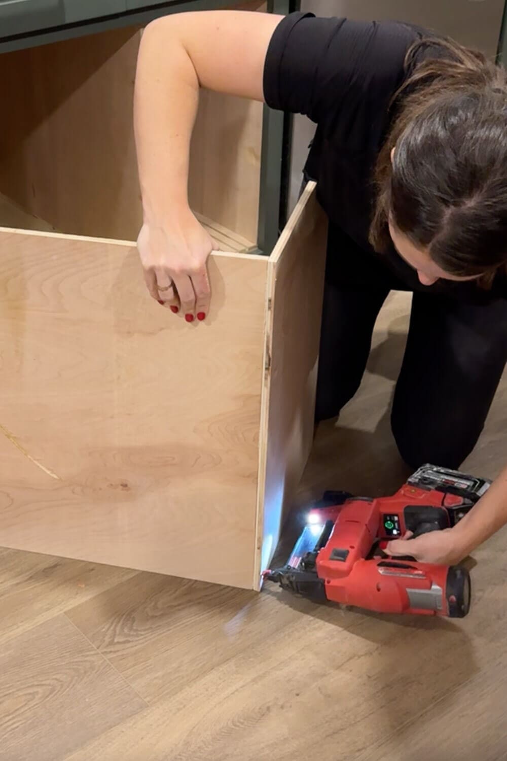 Woman using a cordless brad nailer to attach the side of a plywood drawer box while building a DIY pull-out food storage container organizer for her kitchen cabinet.