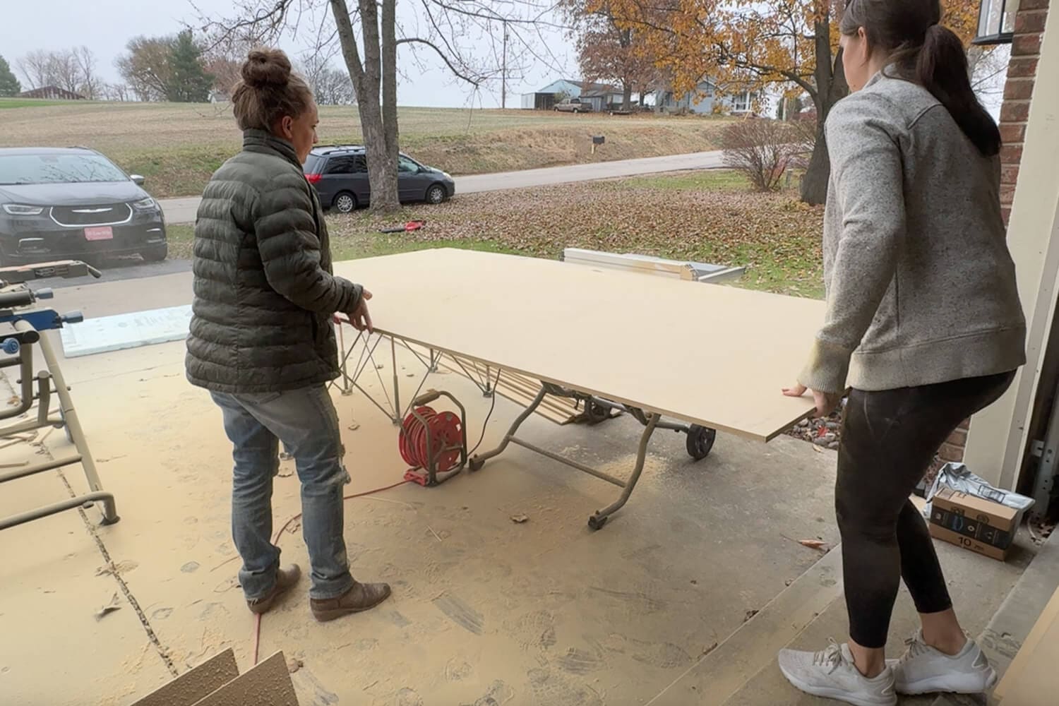 Two women feeding a large sheet of plywood through a table saw in the garage to cut pieces for a DIY vertical storage cabinet insert