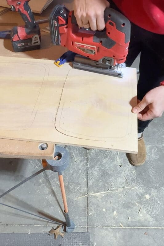 Cutting along the traced outline with a jigsaw to create rounded openings in the plywood top panel for a double trash can pull out cabinet.
