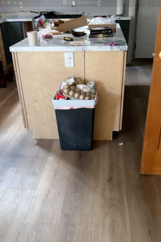 Black kitchen garbage bin overflowing with packaging and food waste sitting in front of an unfinished plywood island, showing the cluttered before view before building a hidden DIY trash can cabinet.
