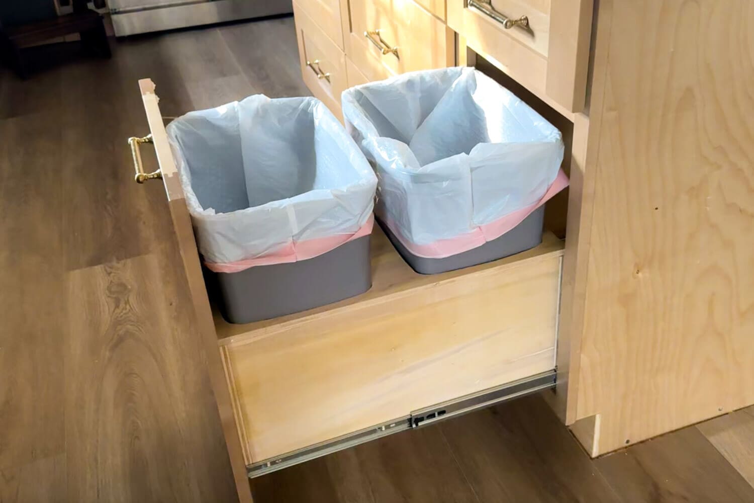 Front view of a DIY double trash can pull out installed in a kitchen cabinet, featuring two lined gray trash bins on a smooth plywood drawer box with side-mounted full-extension slides.