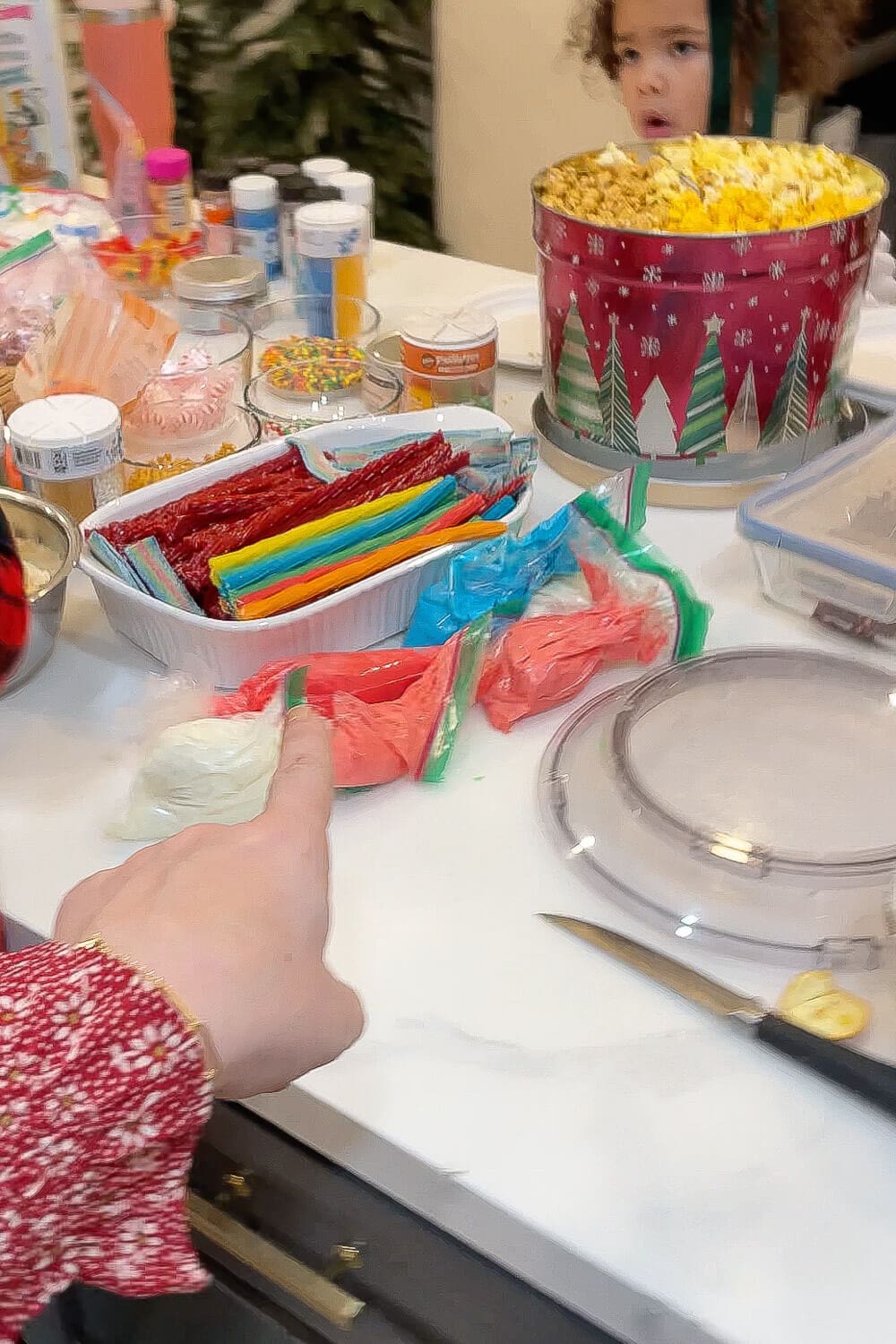 Close up of a gingerbread house candy station with piping bags of red, blue, green, and white royal icing, trays of licorice and rainbow candy strips, sprinkles, and a Christmas popcorn tin.