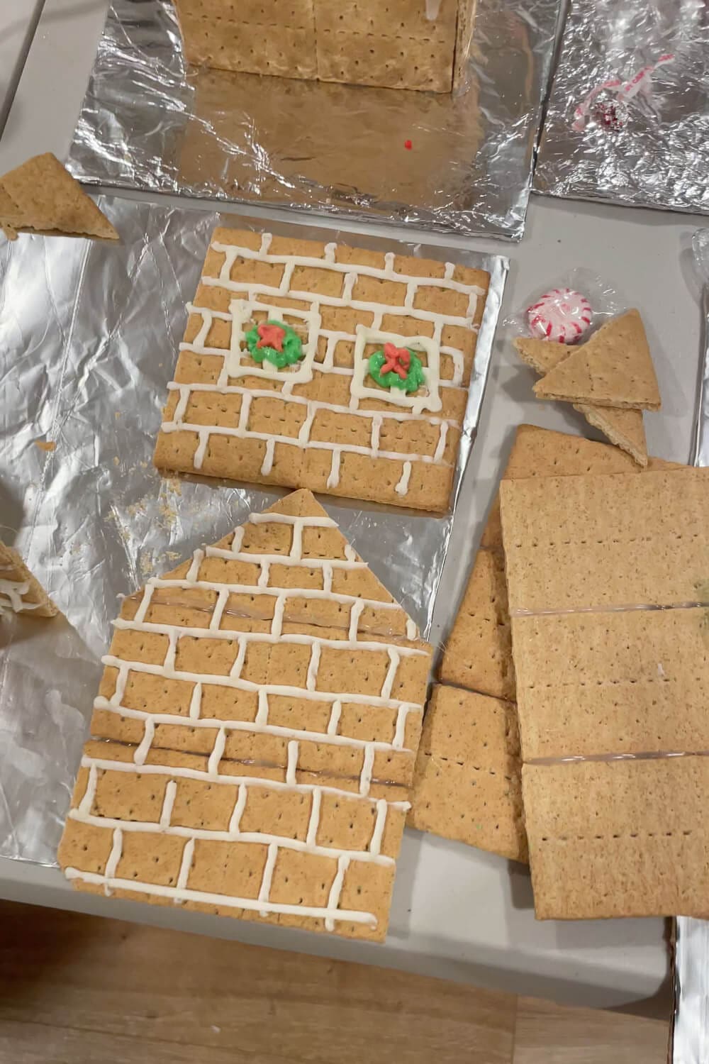 Overhead view of graham cracker gingerbread house panels on foil, outlined in white royal icing with brick lines and wreath windows, with extra cracker pieces and a peppermint candy on the side.