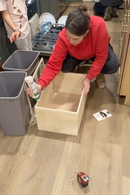 Applying wood glue along the edge of a plywood box to attach the top frame, assembling the pull out trash can cabinet insert next to two gray trash bins.