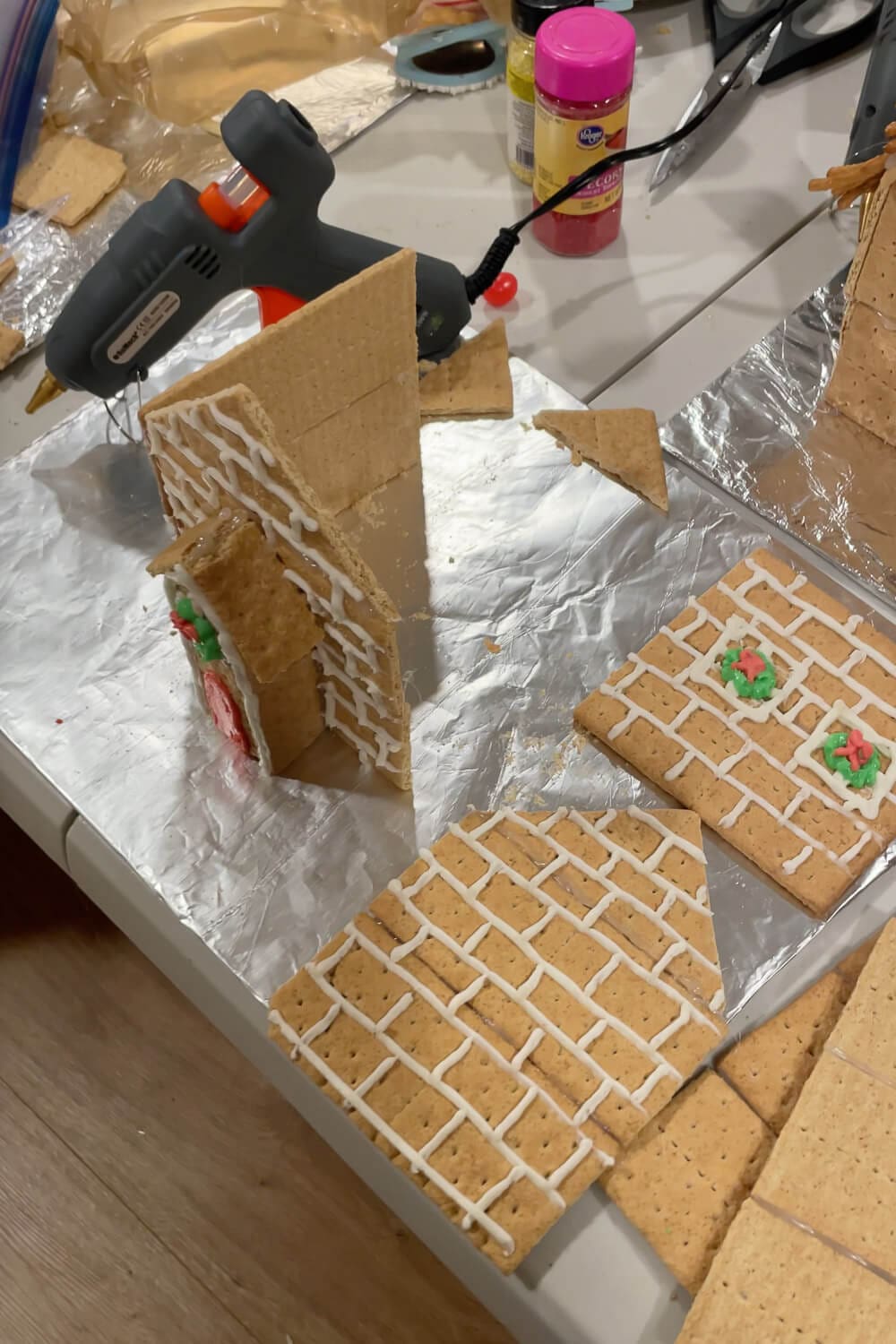 Partially assembled graham cracker gingerbread house on a foil-covered tray, with brick-pattern royal icing on the walls, a hot glue gun nearby, and extra decorated house panels laid flat for decorating.