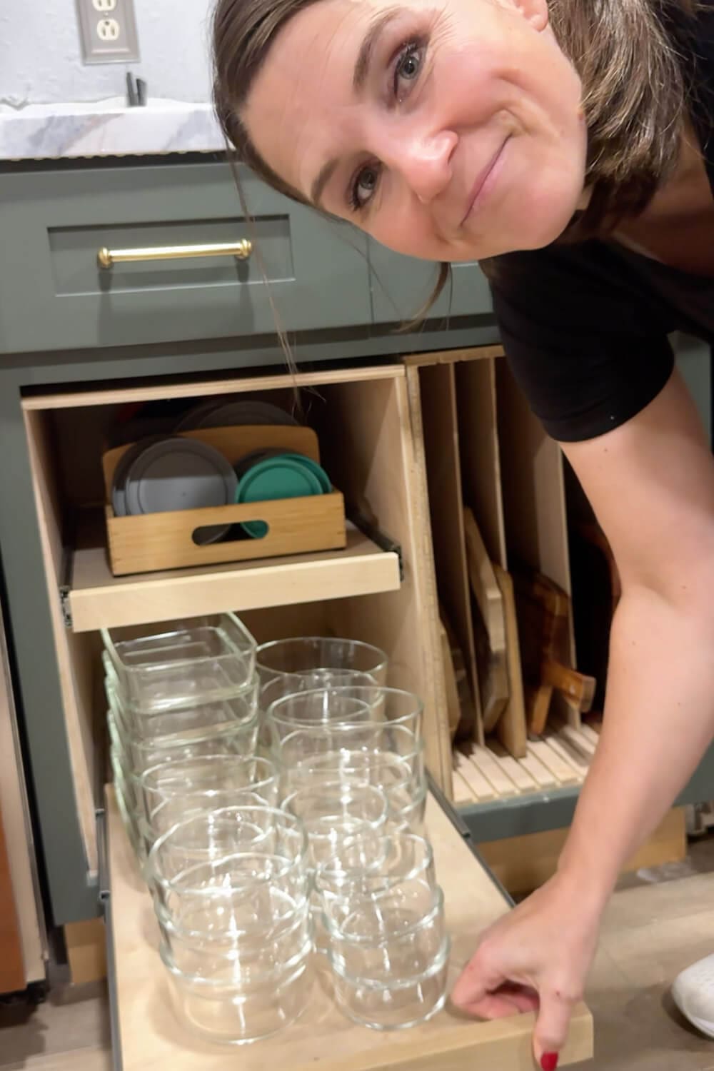 Woman smiling and pulling out a custom drawer organizer filled with neatly stacked glass food storage Tupperware containers and lids in a green kitchen cabinet.