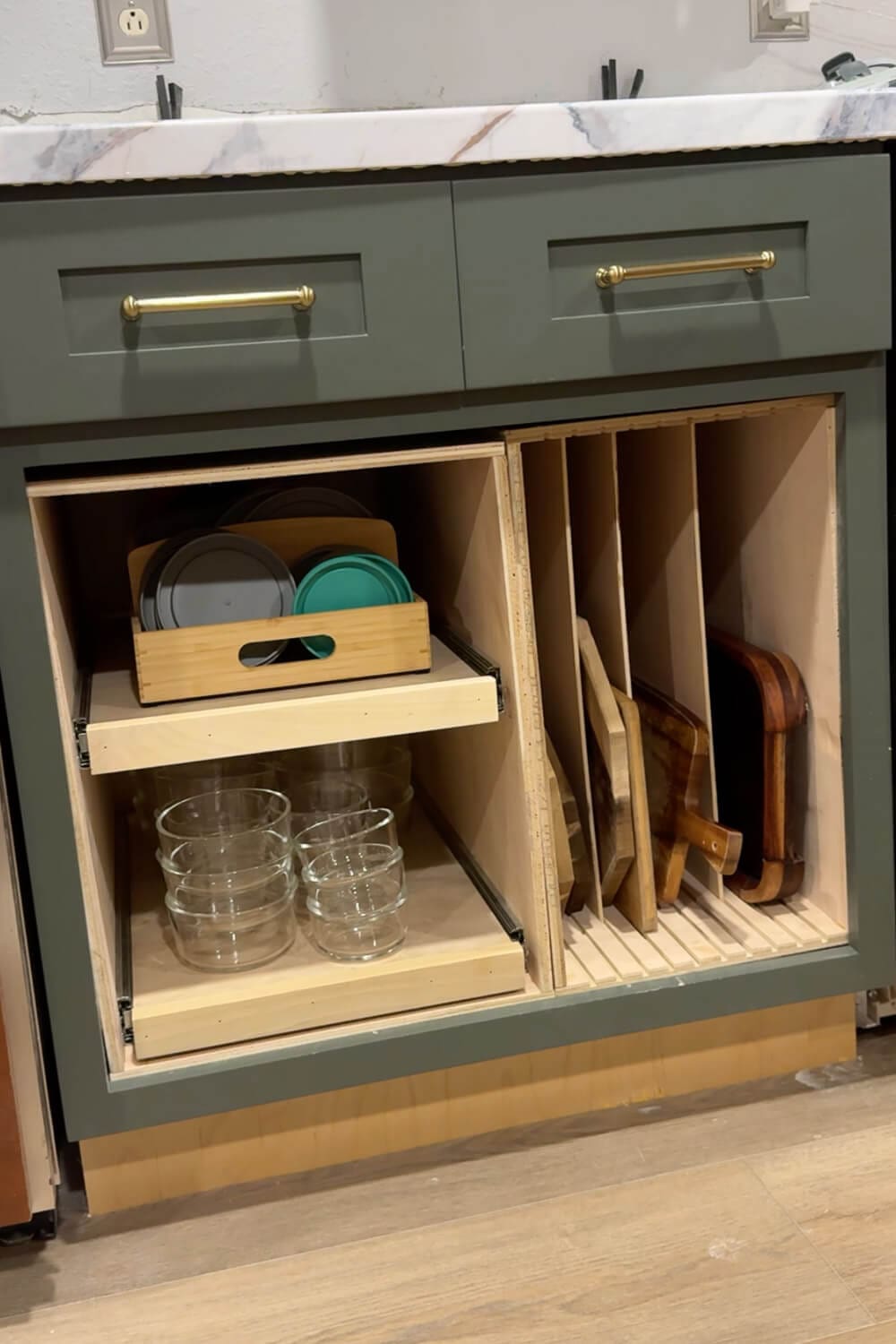 DIY lower kitchen cabinet organizer with glass food storage containers on a pull out shelf and a bamboo bin of lids above, next to vertical cutting board storage in a green cabinet.