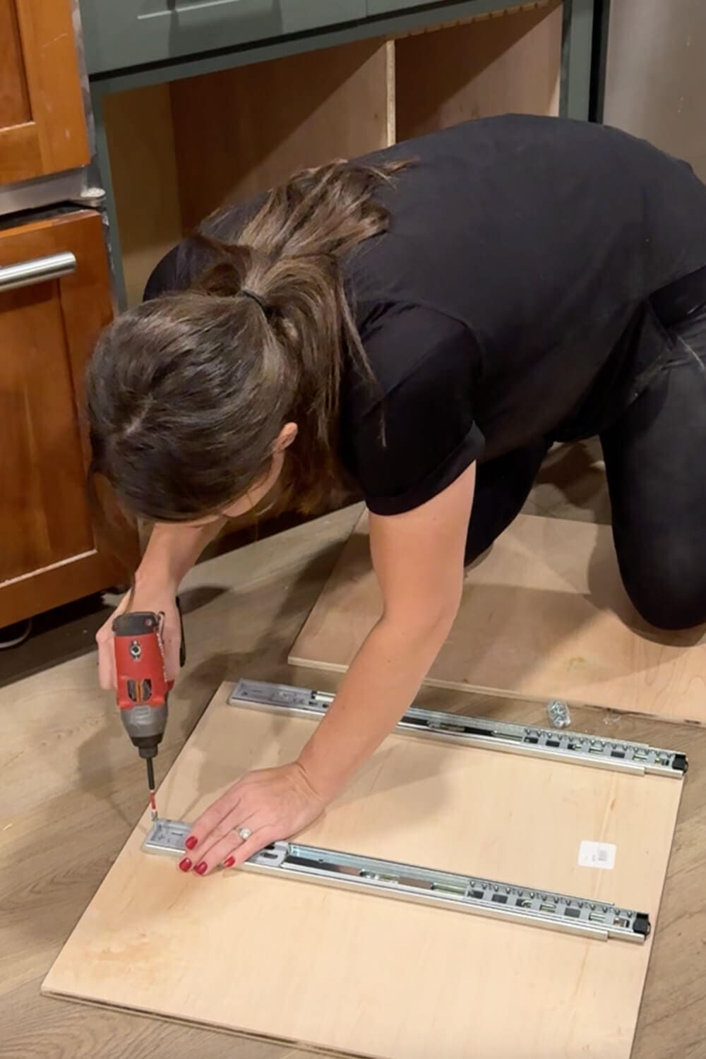 Woman kneeling on the kitchen floor using a drill to attach full-extension drawer slides to a plywood panel for a DIY food storage container organizer.