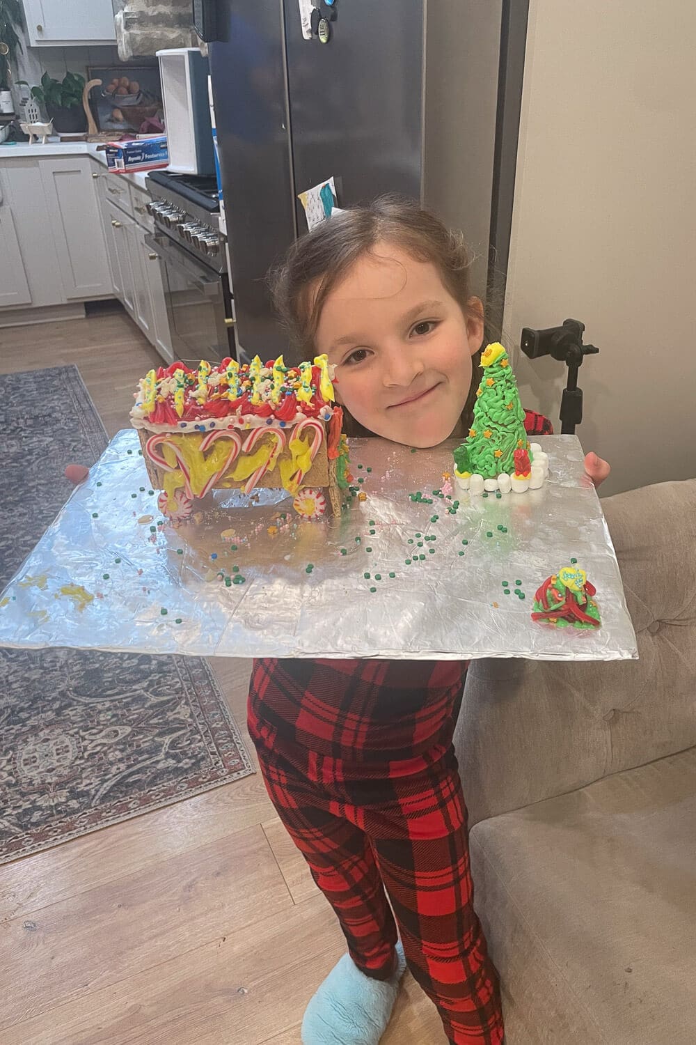 Little girl in red plaid pajamas proudly holding a foil-covered board with her brightly decorated graham cracker gingerbread house and icing Christmas tree.