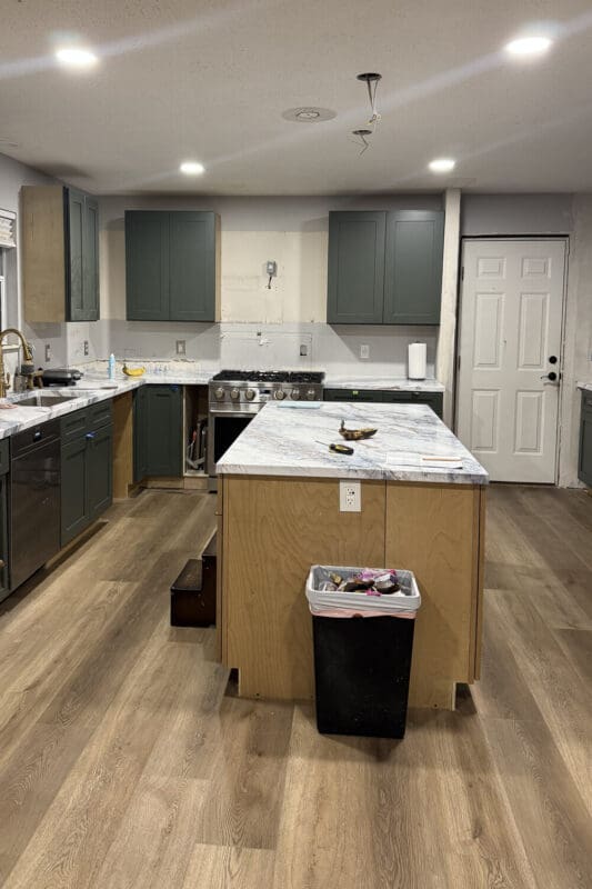 Wide view of a partially finished kitchen with green cabinetry, marble-look countertops, and an unfinished plywood island with a black garbage bin in front.
