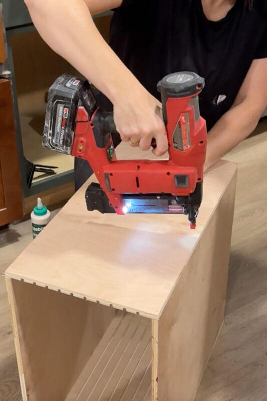 Securing the top panel of a plywood sheet pan organizer box with a brad nailer for a custom kitchen cabinet insert