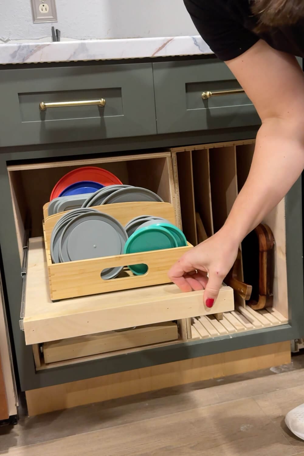 Hand pulling out a DIY slide out shelf holding a bamboo bin filled with colorful food storage Tupperware style container lids next to a vertical cutting board organizer in a lower kitchen cabinet.