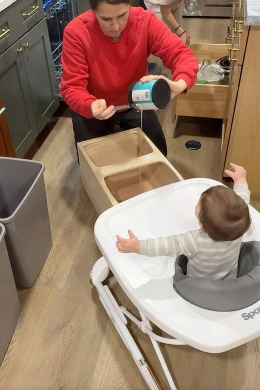 Pouring clear polycrylic into the openings of a plywood garbage can insert while a baby watches from a walker, sealing the wood for a DIY garbage bin drawer.
